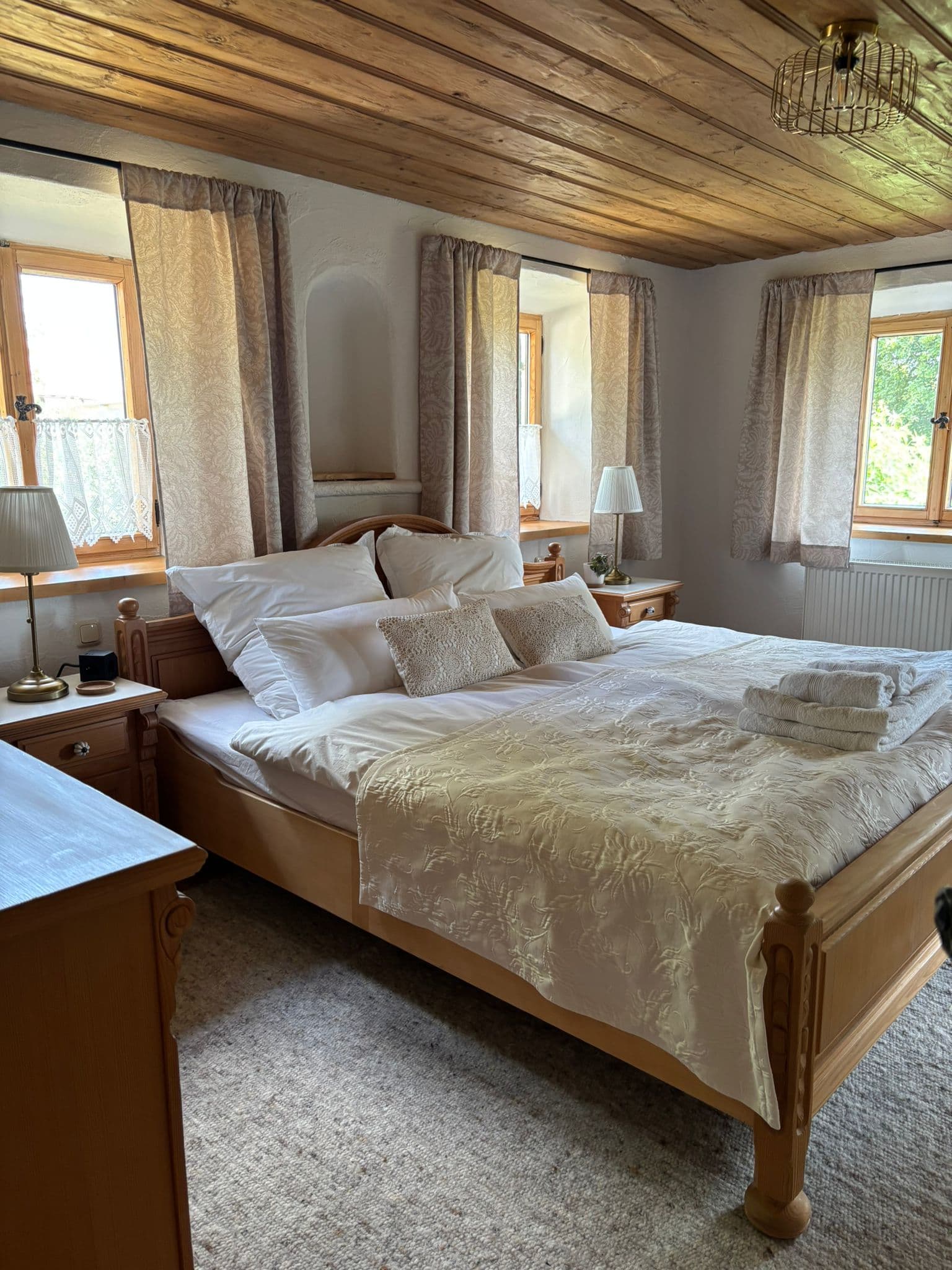 Cozy rustic bedroom with a wooden bed, white linens, patterned curtains, and a wood-paneled ceiling.